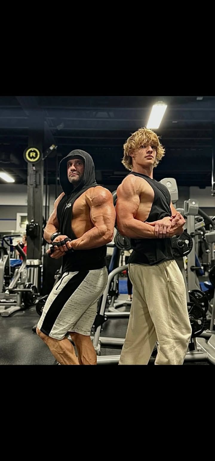 Two muscular men posing back-to-back in a gym, wearing hoodies and workout pants.
