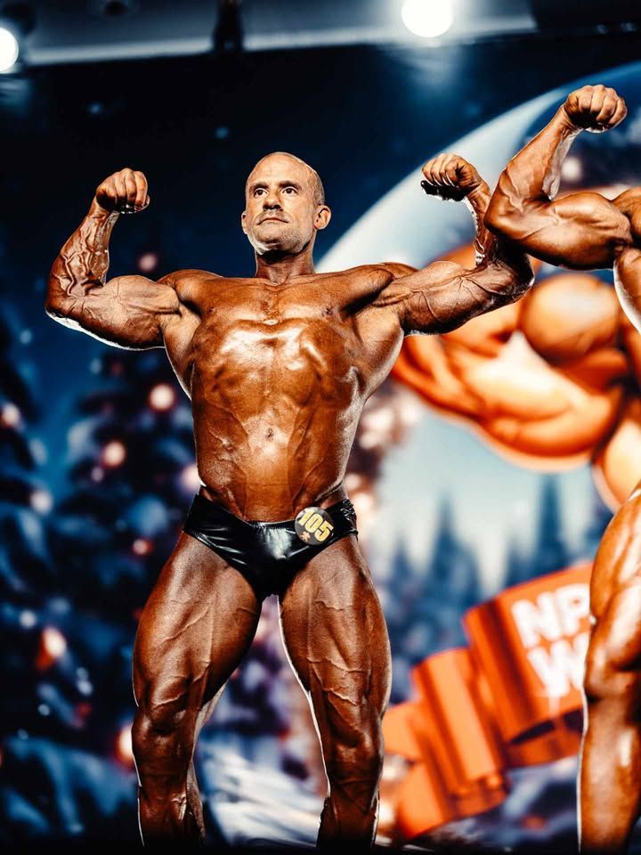 Bodybuilder posing onstage in black trunks, flexing under bright lights with blurred crowd in the background.
