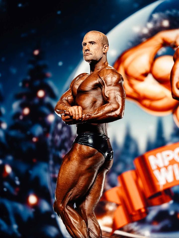 Bodybuilder posing on stage in black trunks, with a blurred competition backdrop and bright lights.