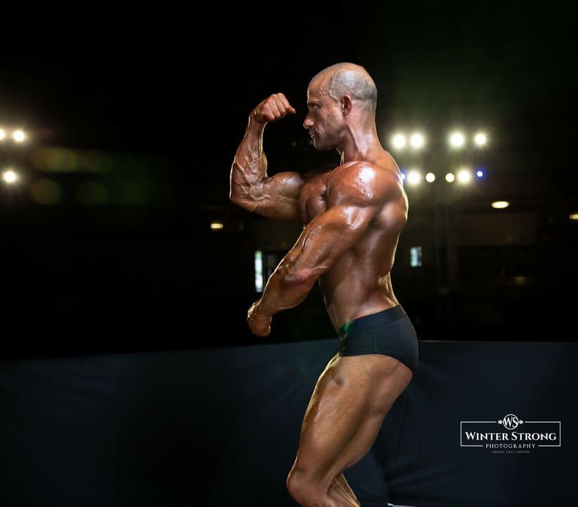 Bodybuilder posing on stage under bright spotlights, flexing a side bicep in black trunks.