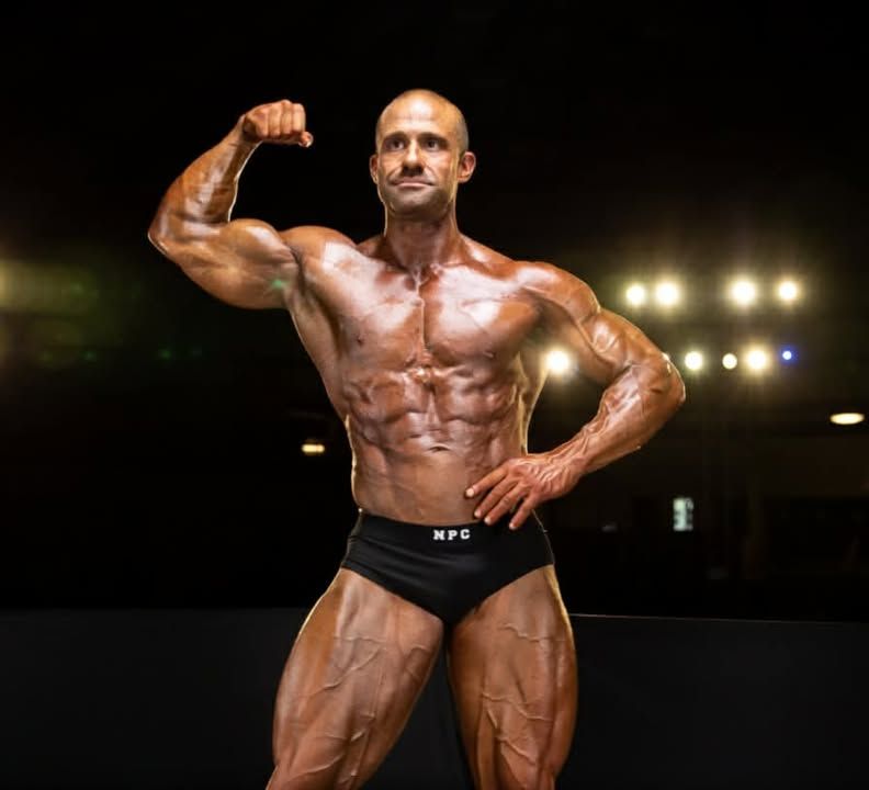 Bodybuilder flexing on a stage under bright lights, wearing black trunks.