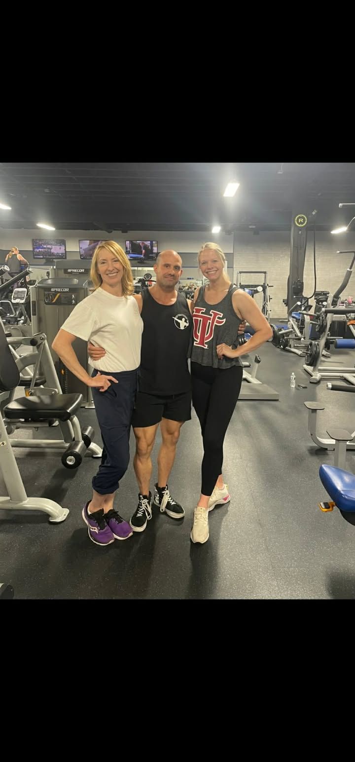 Three people posing together in a gym, smiling under bright lights, with exercise equipment in the background.