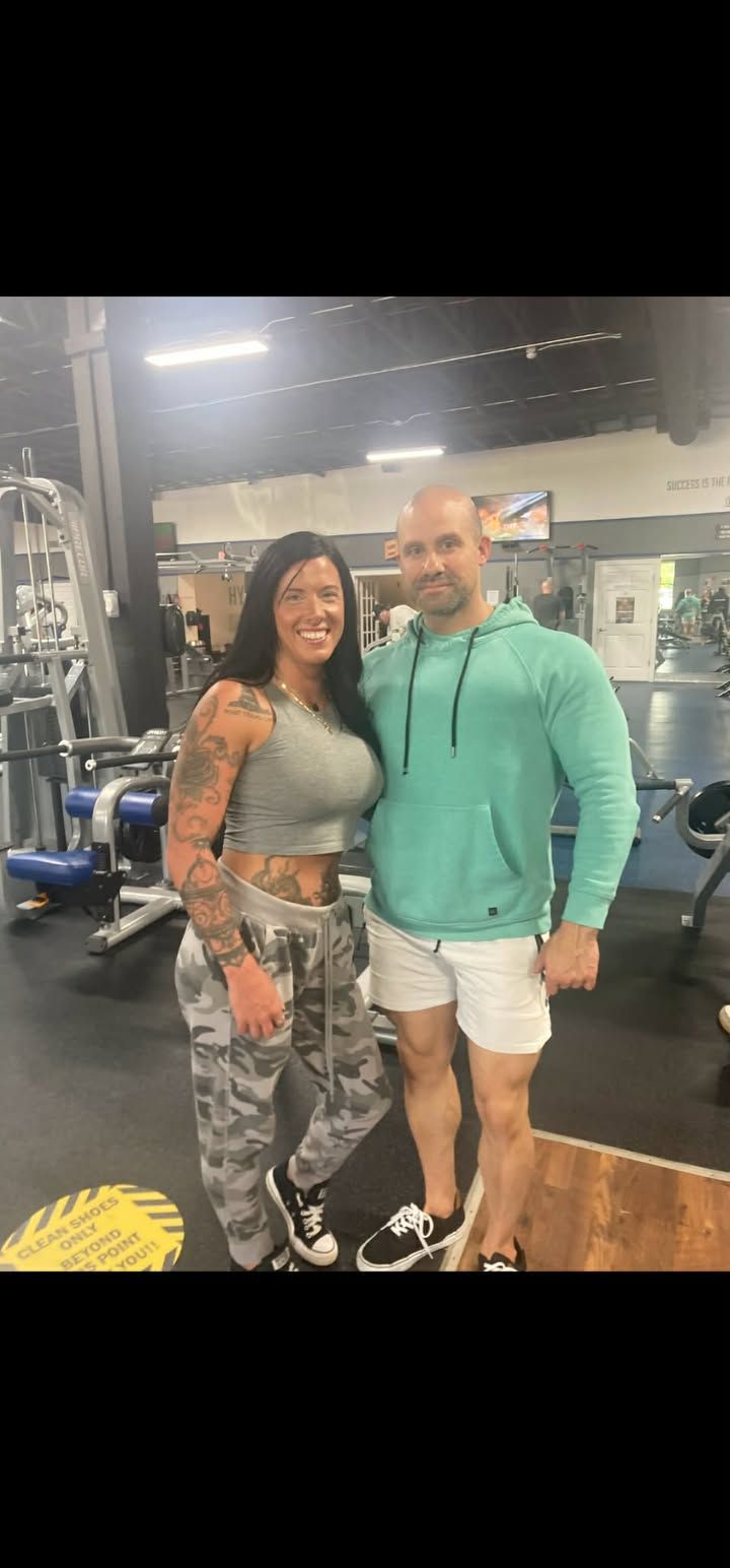 Two people smiling in a gym, standing together in workout clothes near weight equipment.