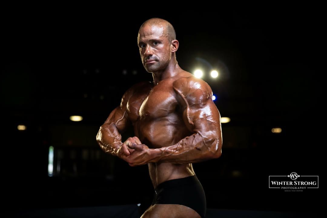 Bodybuilder posing on stage under dramatic lights, wearing black trunks and oil-sheened muscles.