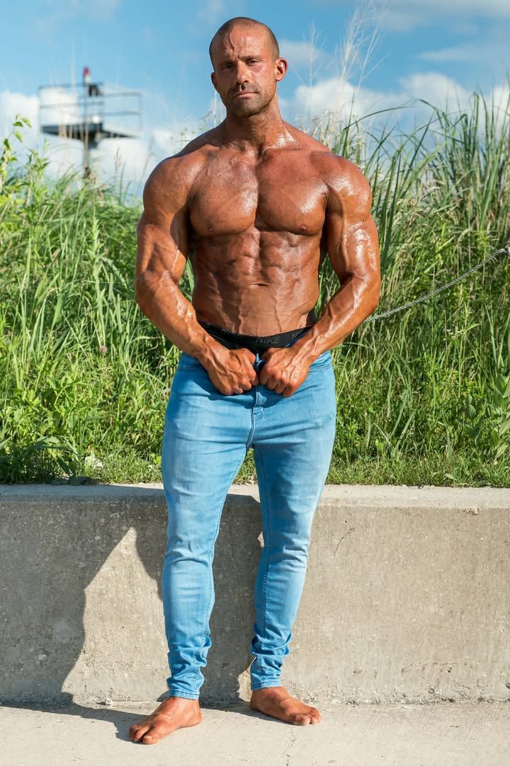 Muscular shirtless man posing outdoors in jeans, standing by a concrete wall with tall grass behind him