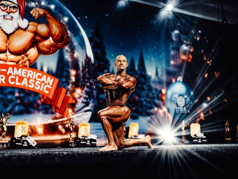 Bodybuilder posing on a lit stage with an American Classic bodybuilding backdrop