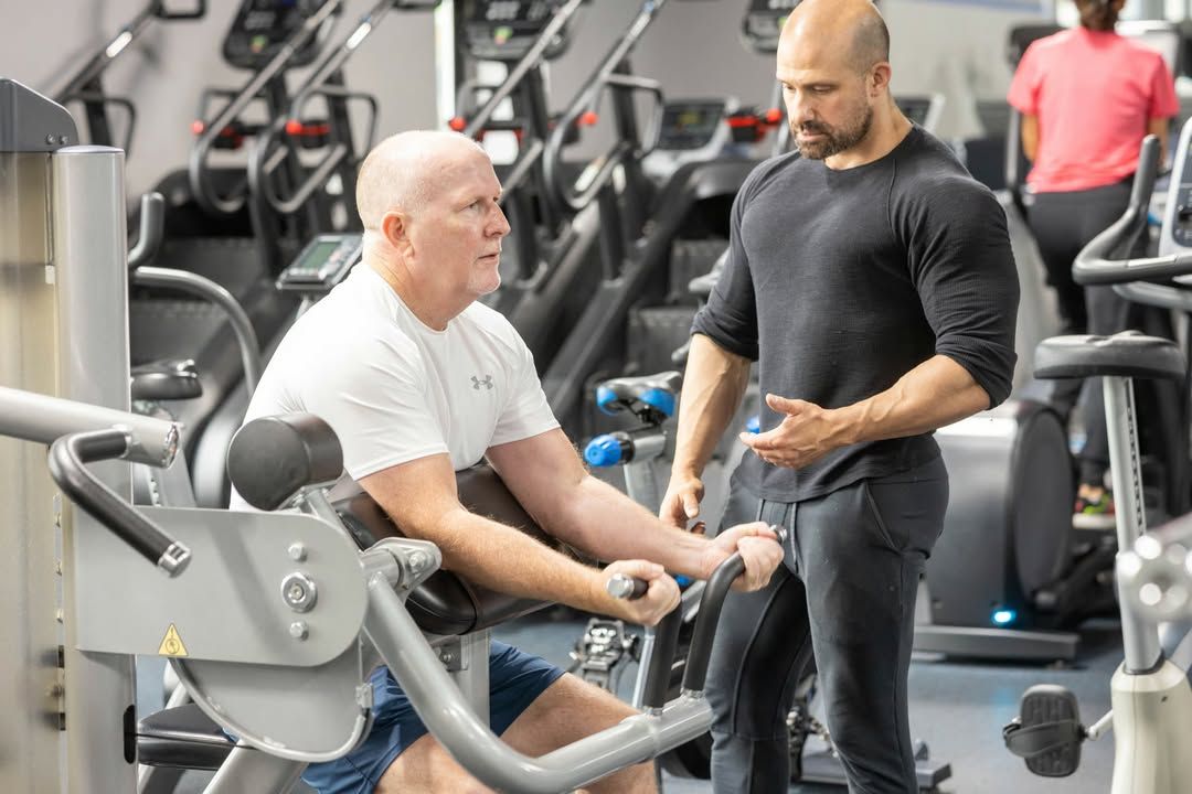 Two men in a gym, one seated on equipment and one standing beside him talking.
