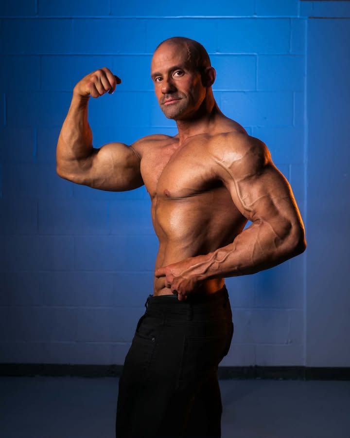 Muscular shirtless man flexing his arm against a blue wall in a gym-like setting