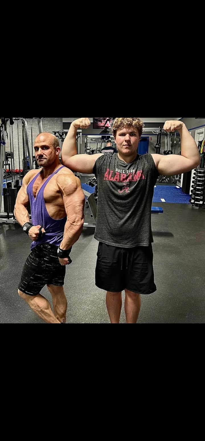 Two men posing in a gym, flexing their arms in workout clothes.