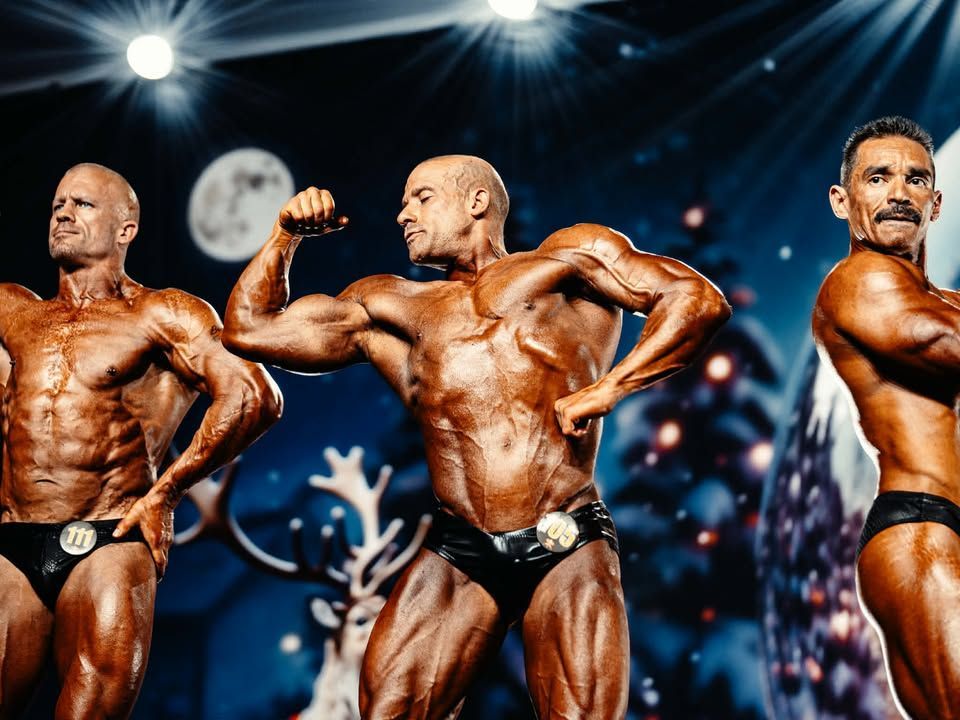Three male bodybuilders on stage under bright spotlights, posing in black trunks during a competition.