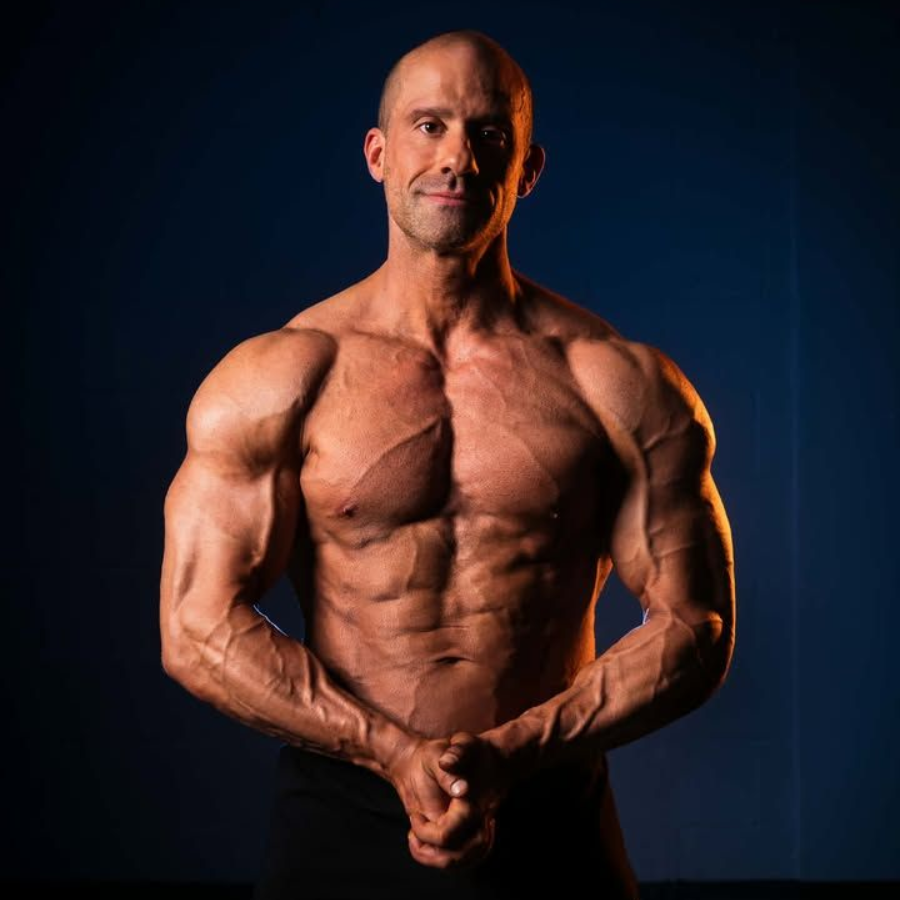 Muscular bald man posing front-facing against a dark blue background, hands clasped at waist