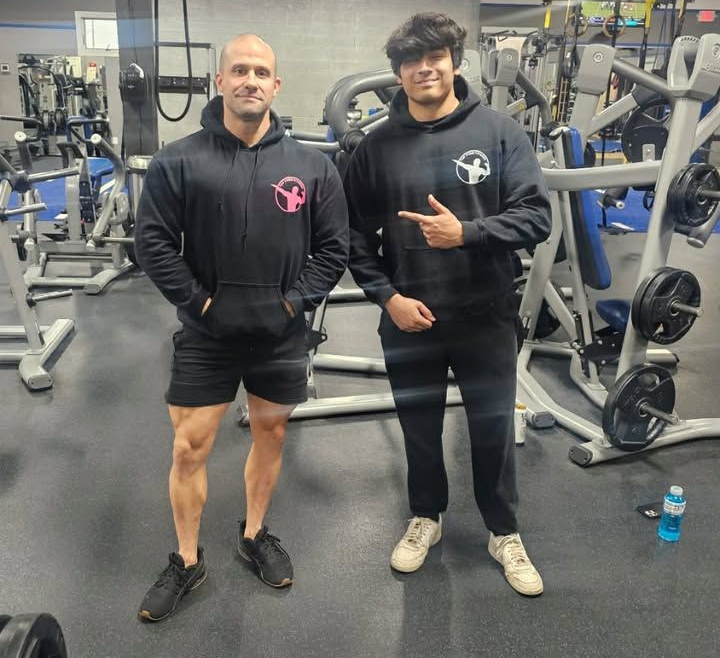 Two men posing in a gym, both wearing black hoodies and athletic pants, with exercise equipment behind them.