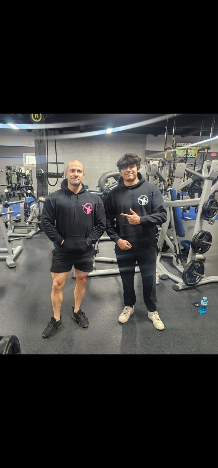 Two people posing in a gym, one flexing, with weight machines in the background.