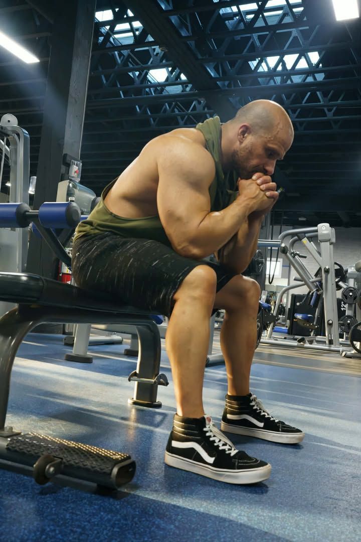Person seated on a gym bench, leaning forward with elbows on knees in a workout area.
