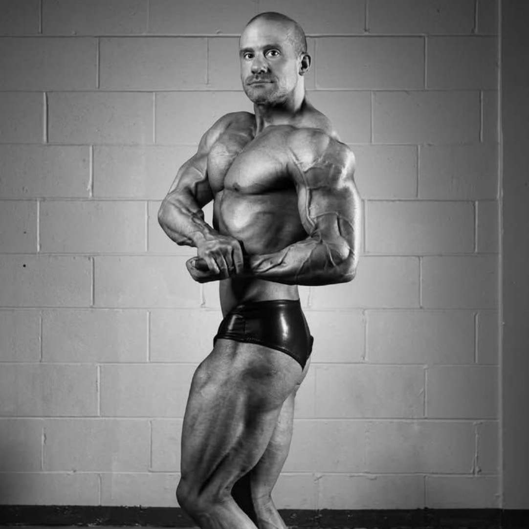 Bodybuilder posing in a gym, flexing muscles in black trunks against a cinderblock wall.