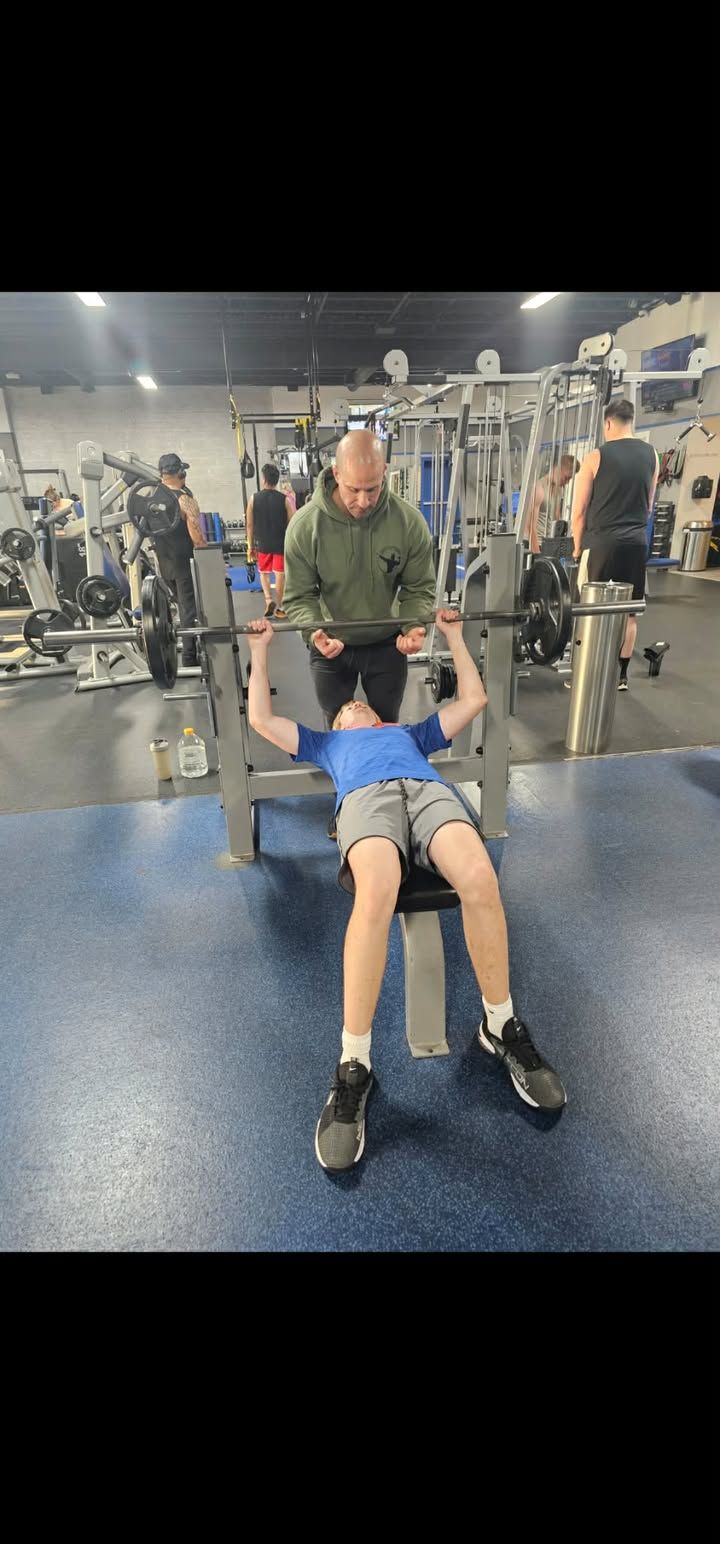 Person bench pressing a barbell in a gym, with a spotter standing behind.