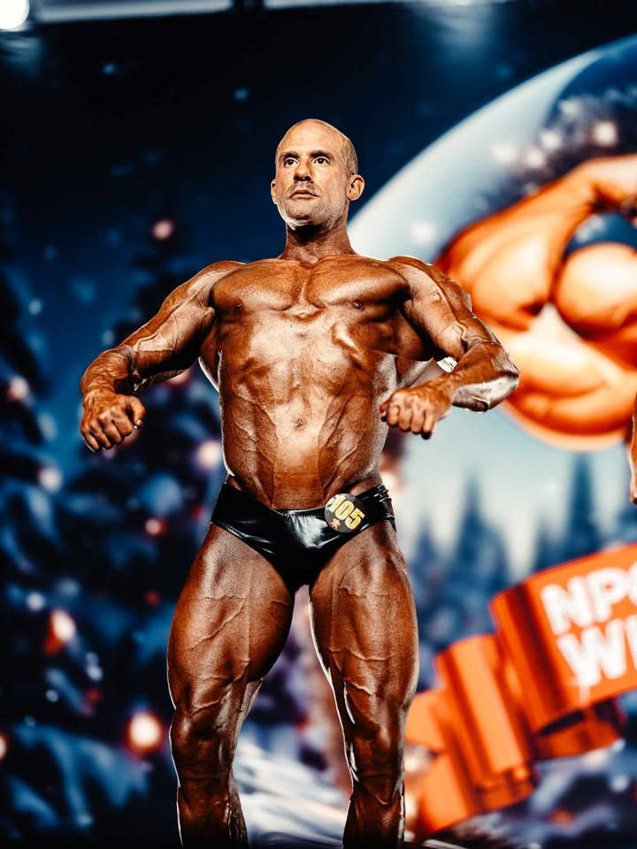 Bodybuilder posing onstage in dark trunks, with a blue and orange competition backdrop.