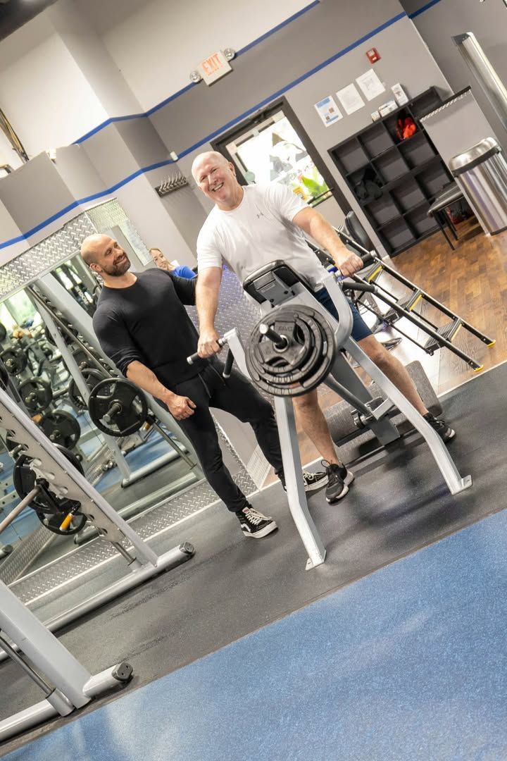 Two people in a gym, one using a weight machine with stacked plates and one standing beside them.