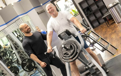 Two men in a gym posing with a weight machine, one in black and one in white