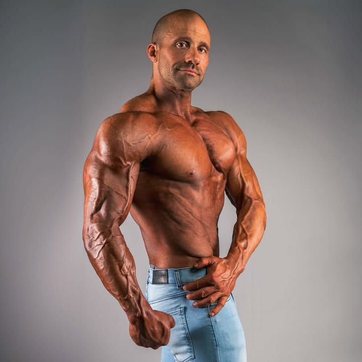Muscular bodybuilder posing shirtless against a gray studio background, wearing light blue jeans.