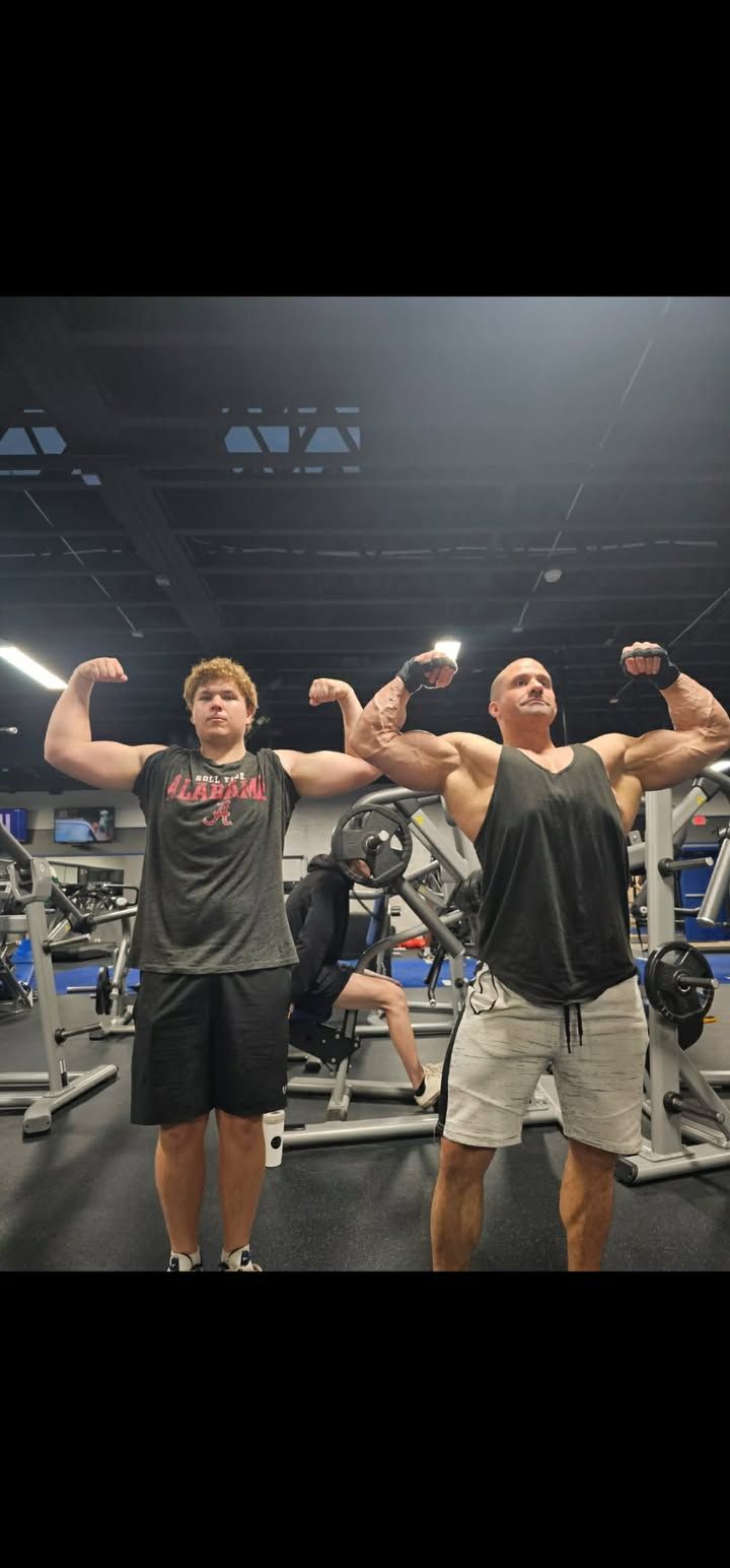 Two people flexing in a gym under bright lights, with exercise machines in the background