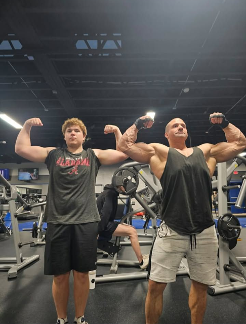 Two people flexing biceps in a gym, one in a black shirt and one in a tank top.