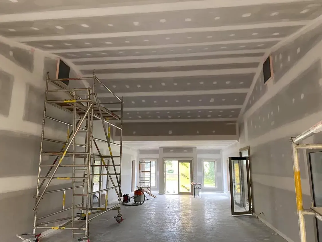 Unfinished room with drywall, scaffolding, and bright light from large windows.