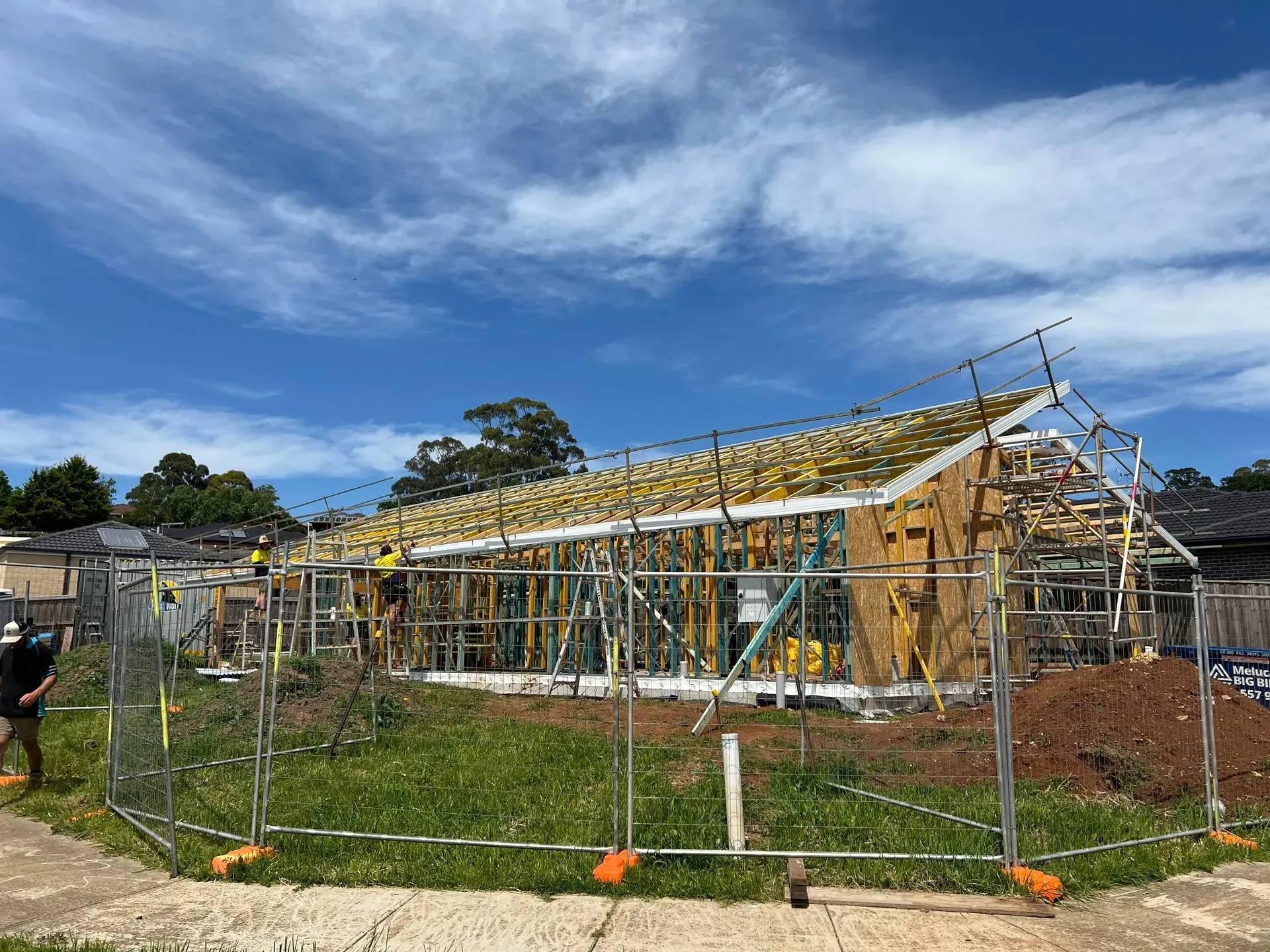 House frame under construction behind a chain-link fence, with wooden roof trusses and blue sky overhead