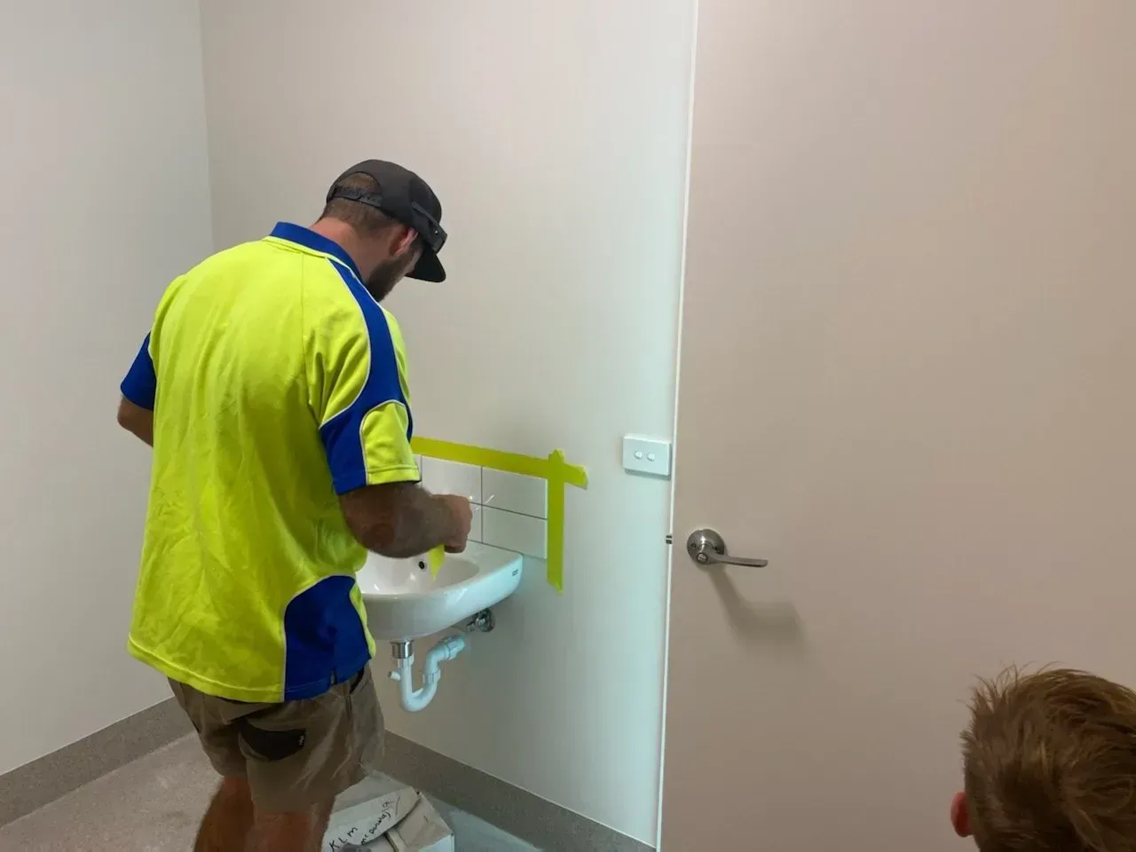 Worker installing a wall-mounted sink in a bathroom, with another person partially visible nearby.