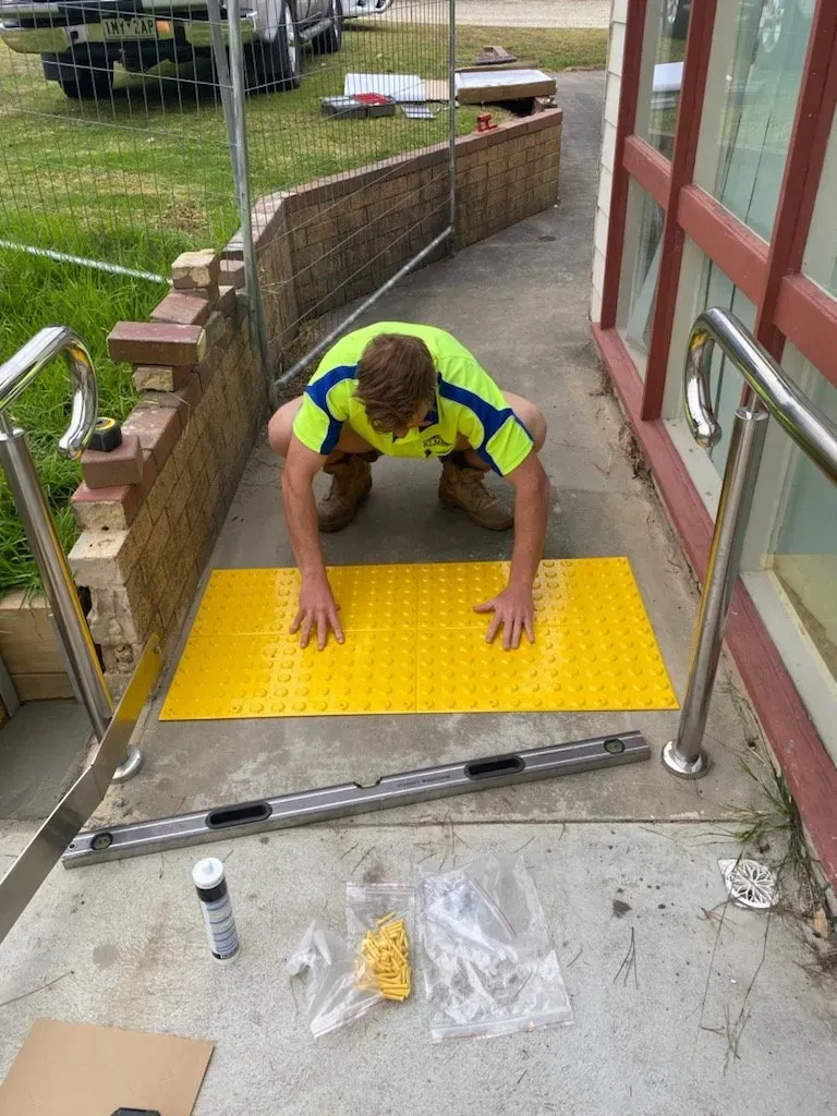 Worker kneeling on a yellow tactile paving panel at a building entrance, installing or inspecting it.