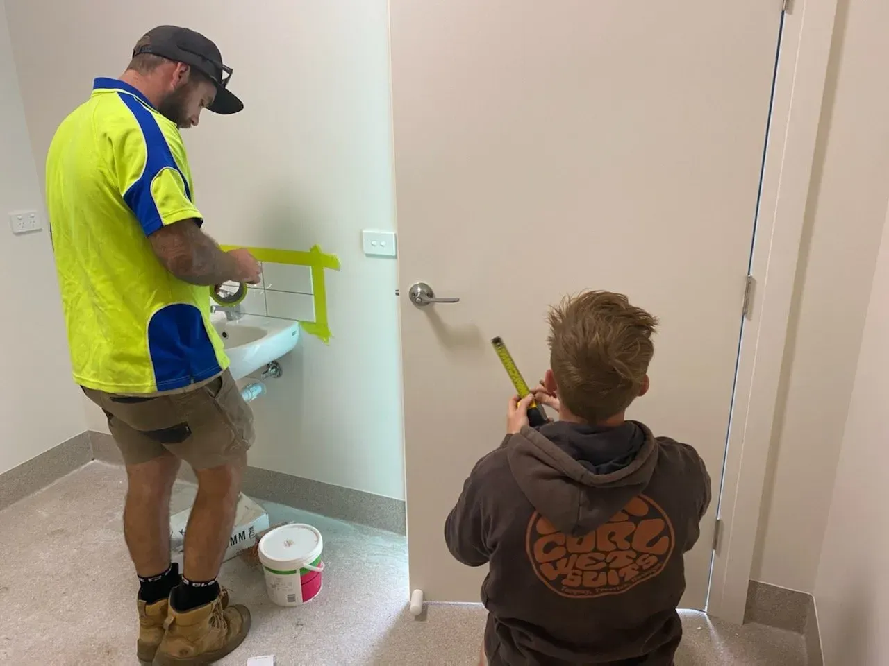 Two people painting a bathroom wall yellow, one standing and one kneeling near a white toilet.