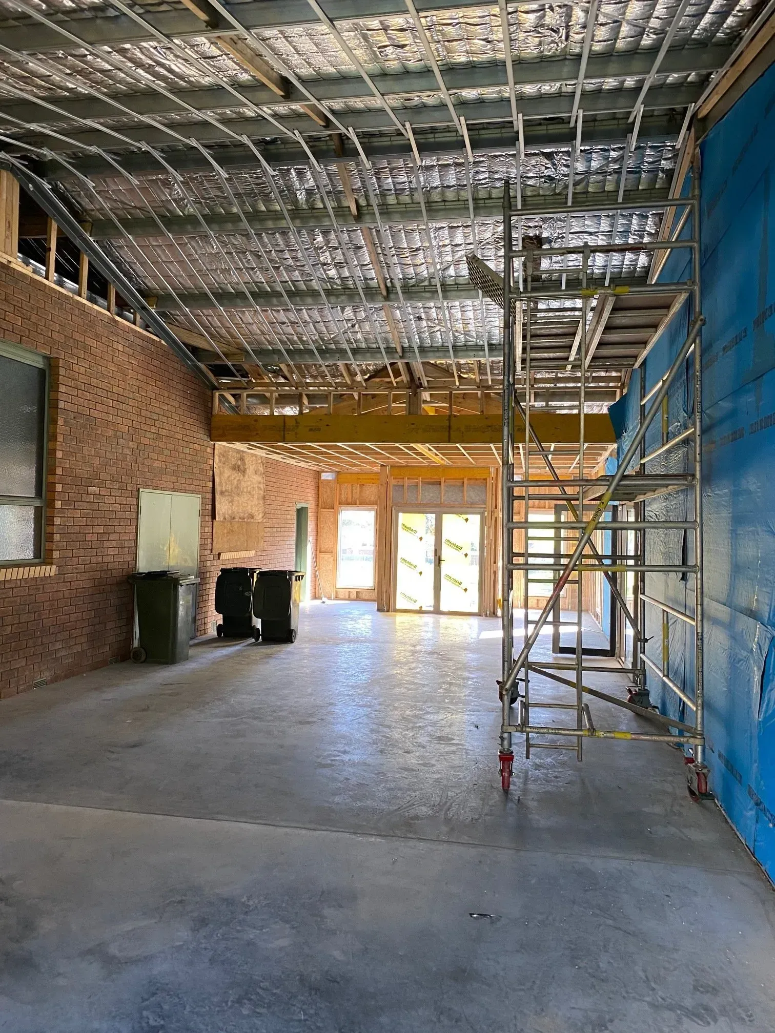 Empty warehouse interior with exposed ceiling, brick wall, blue scaffolding, and sunlight at the far end