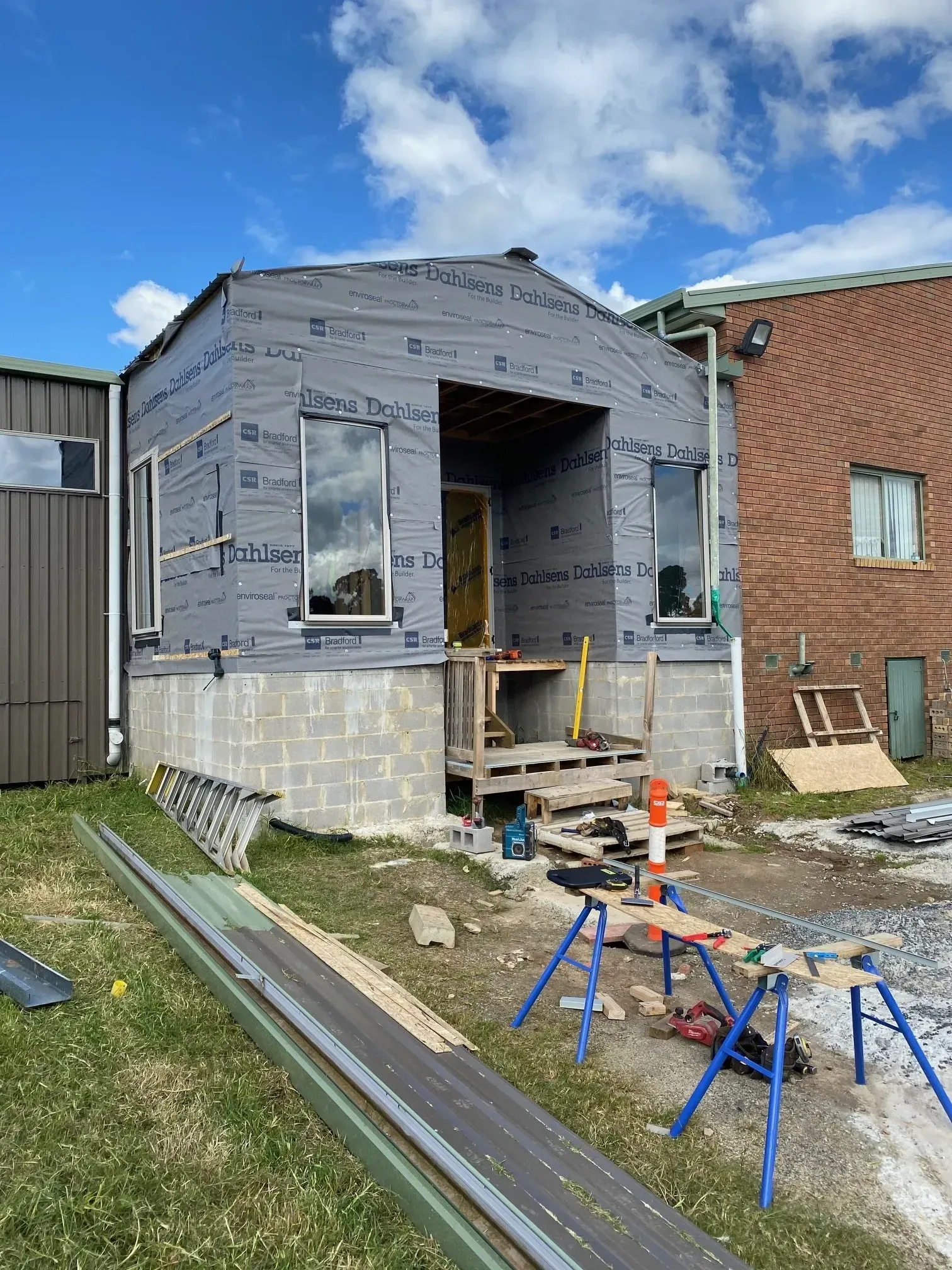 Small house under construction with gray siding, front porch steps, and tools on a grassy lot