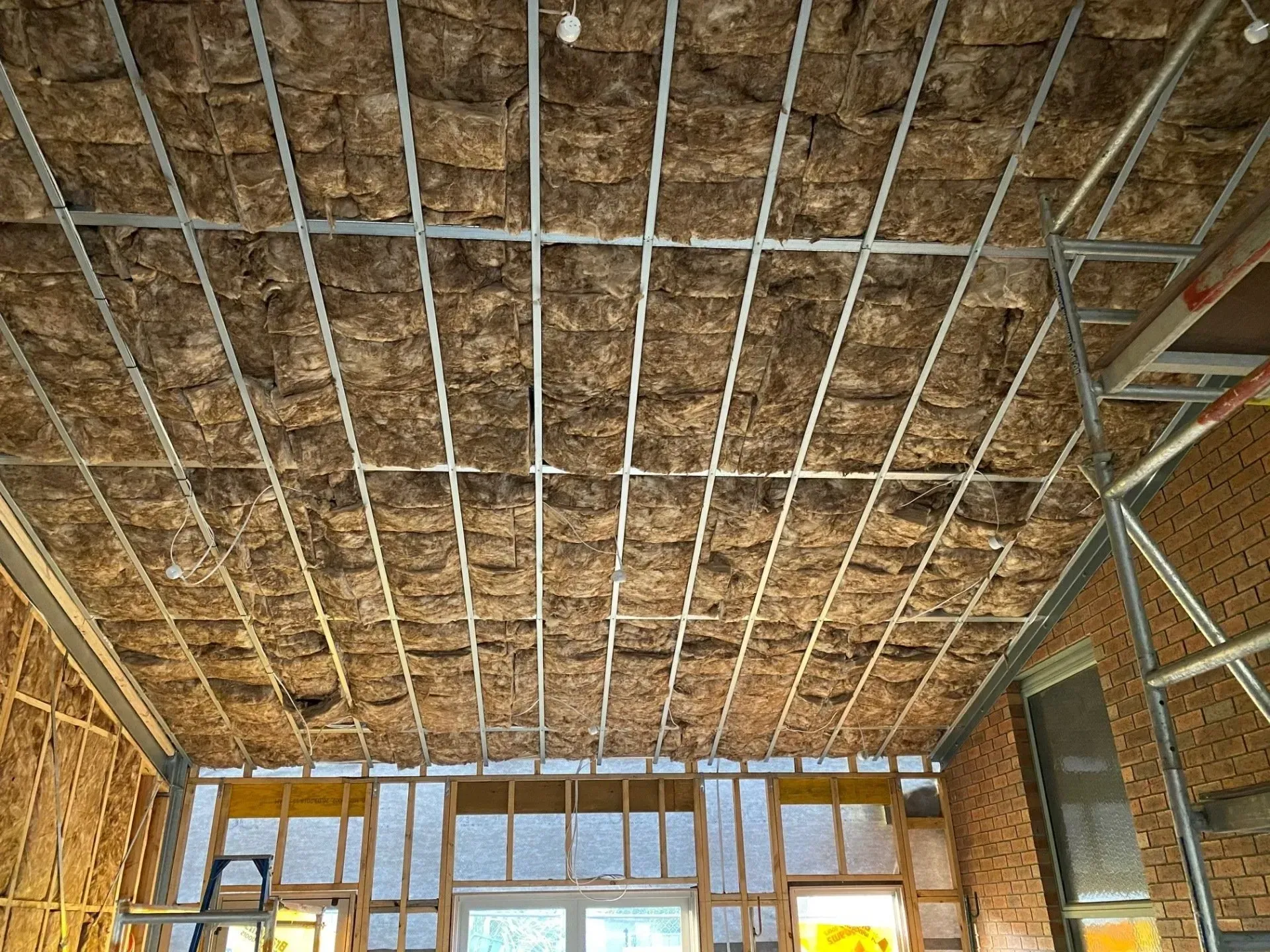 Exposed ceiling framing with insulation in a room under construction, showing windows and brick walls below.