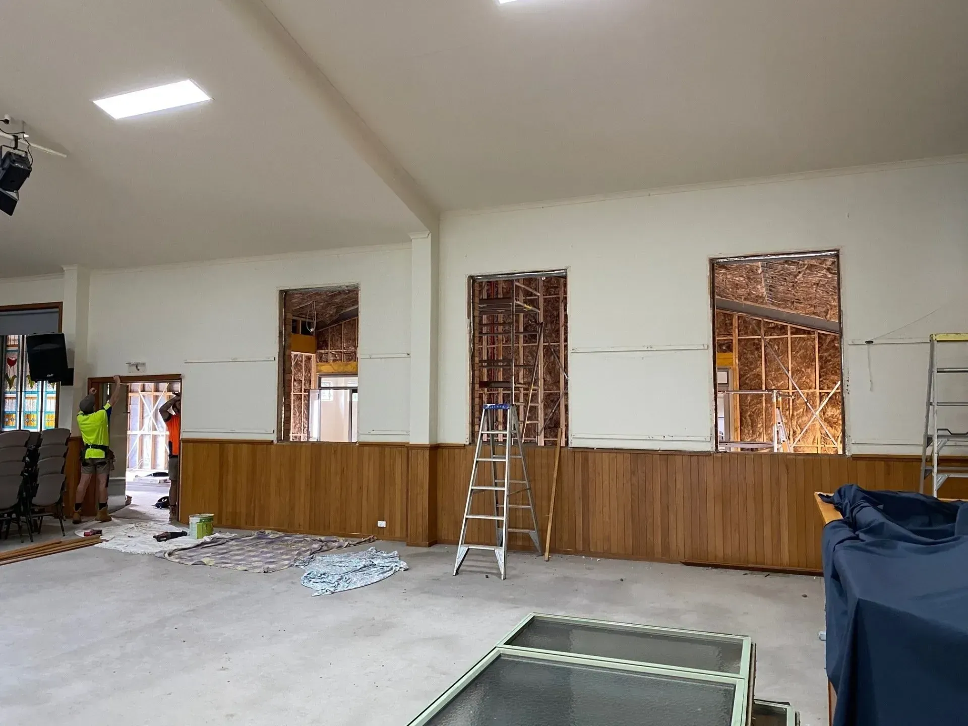 Partially renovated hall with wood paneling, ladders, and exposed windows inside a bright room