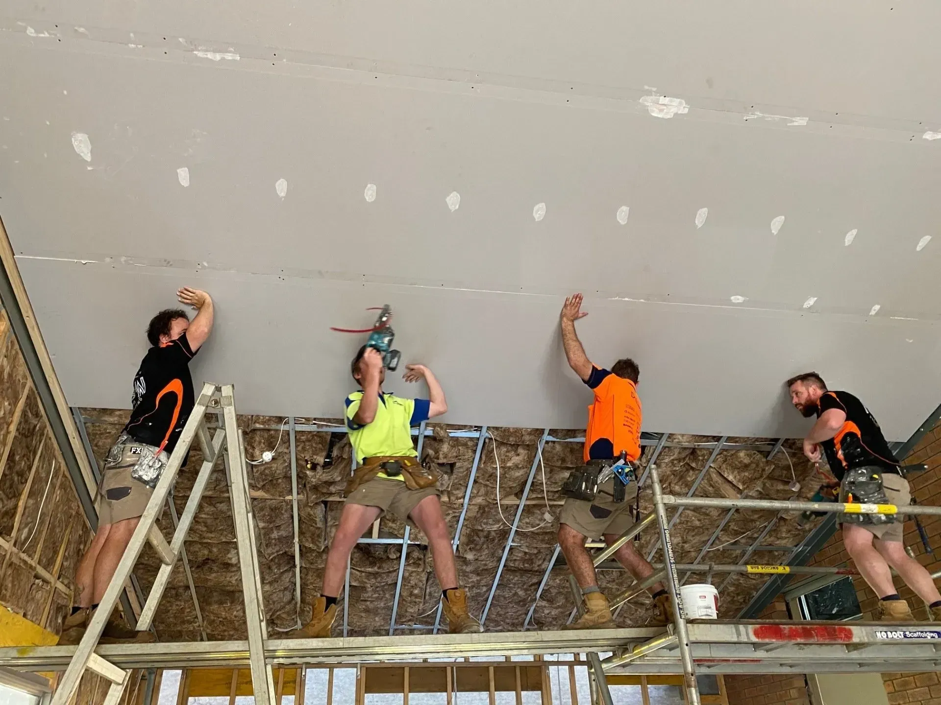 Four workers installing drywall on a ceiling at a construction site, using scaffolding and tools
