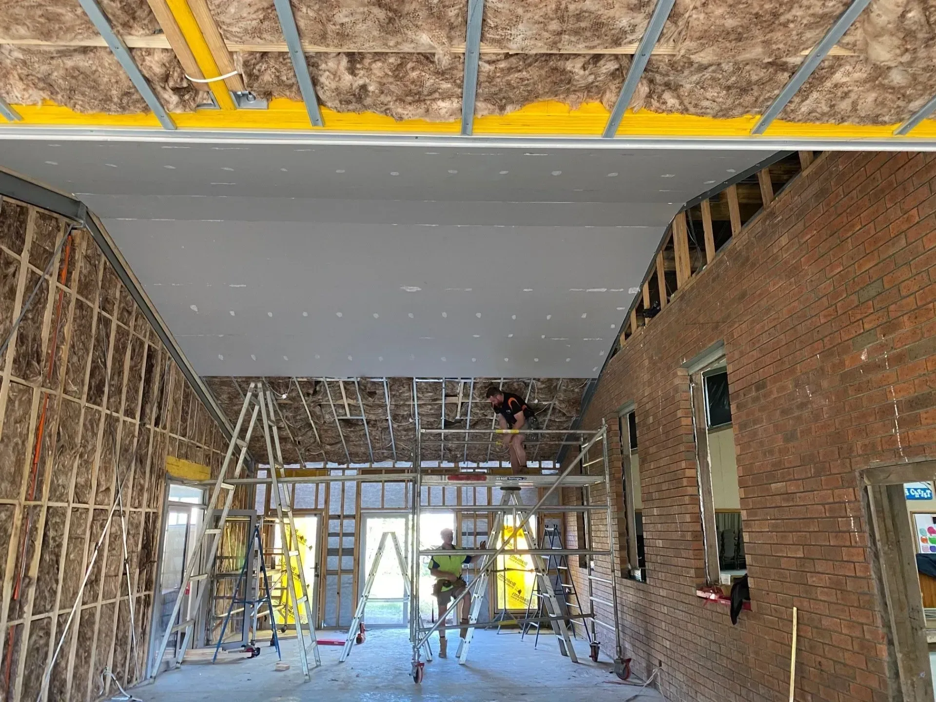 Interior of a building under construction, with scaffolding, drywall, and workers on ladders.