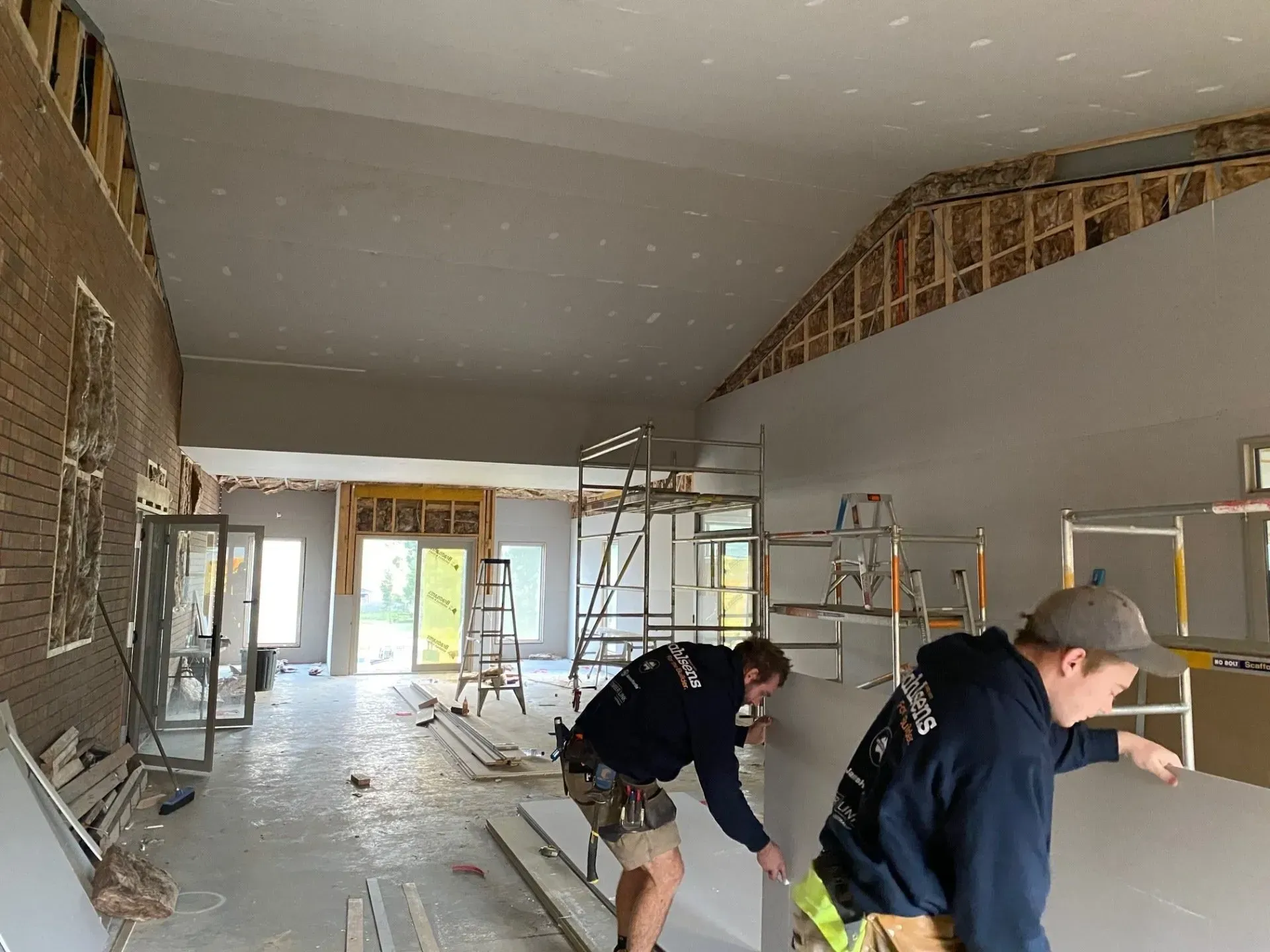Workers sanding a large unfinished room with exposed framing and scaffolding inside.