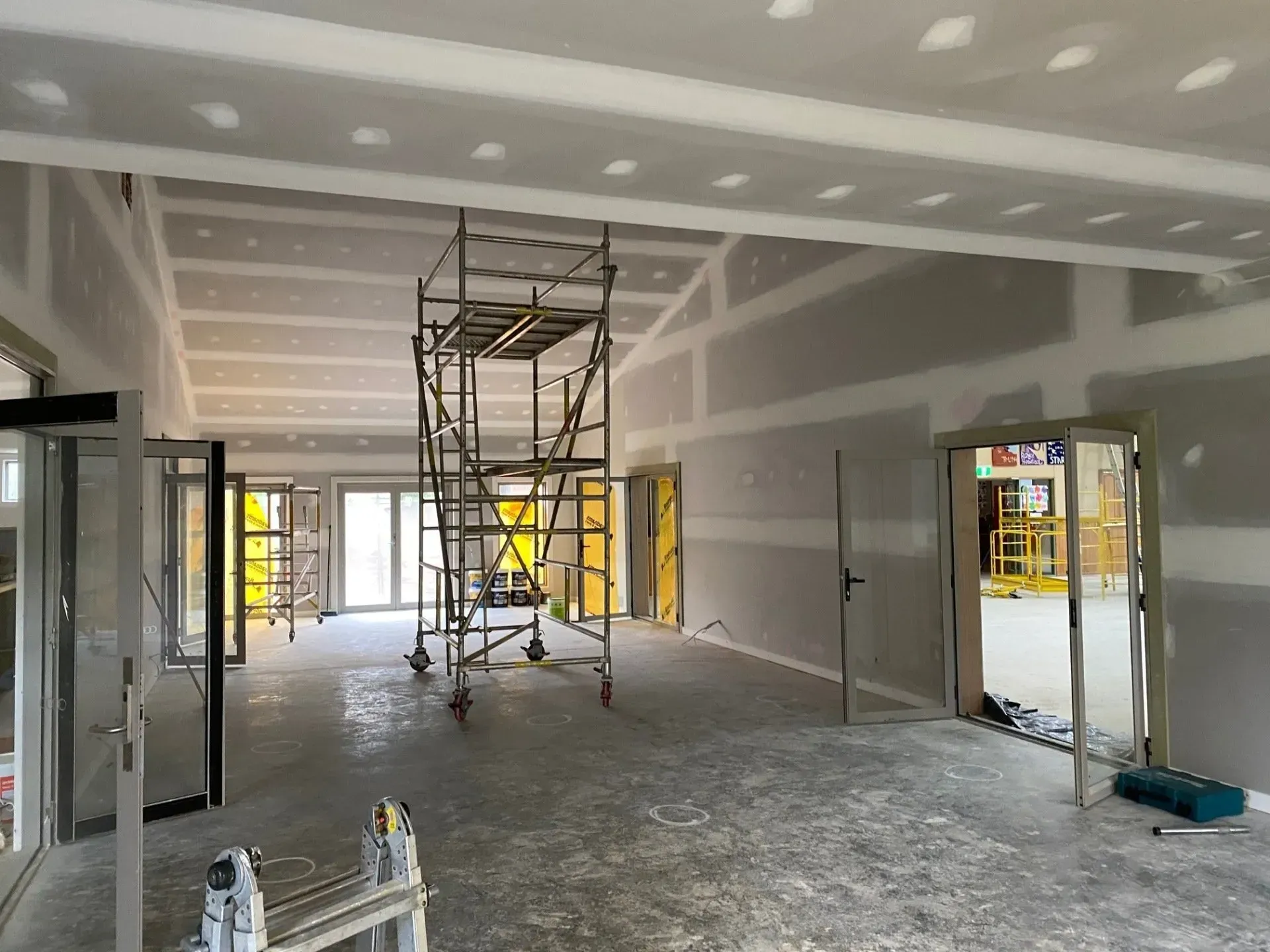 Interior under construction with scaffolding, unfinished drywall, and tools on a concrete floor.