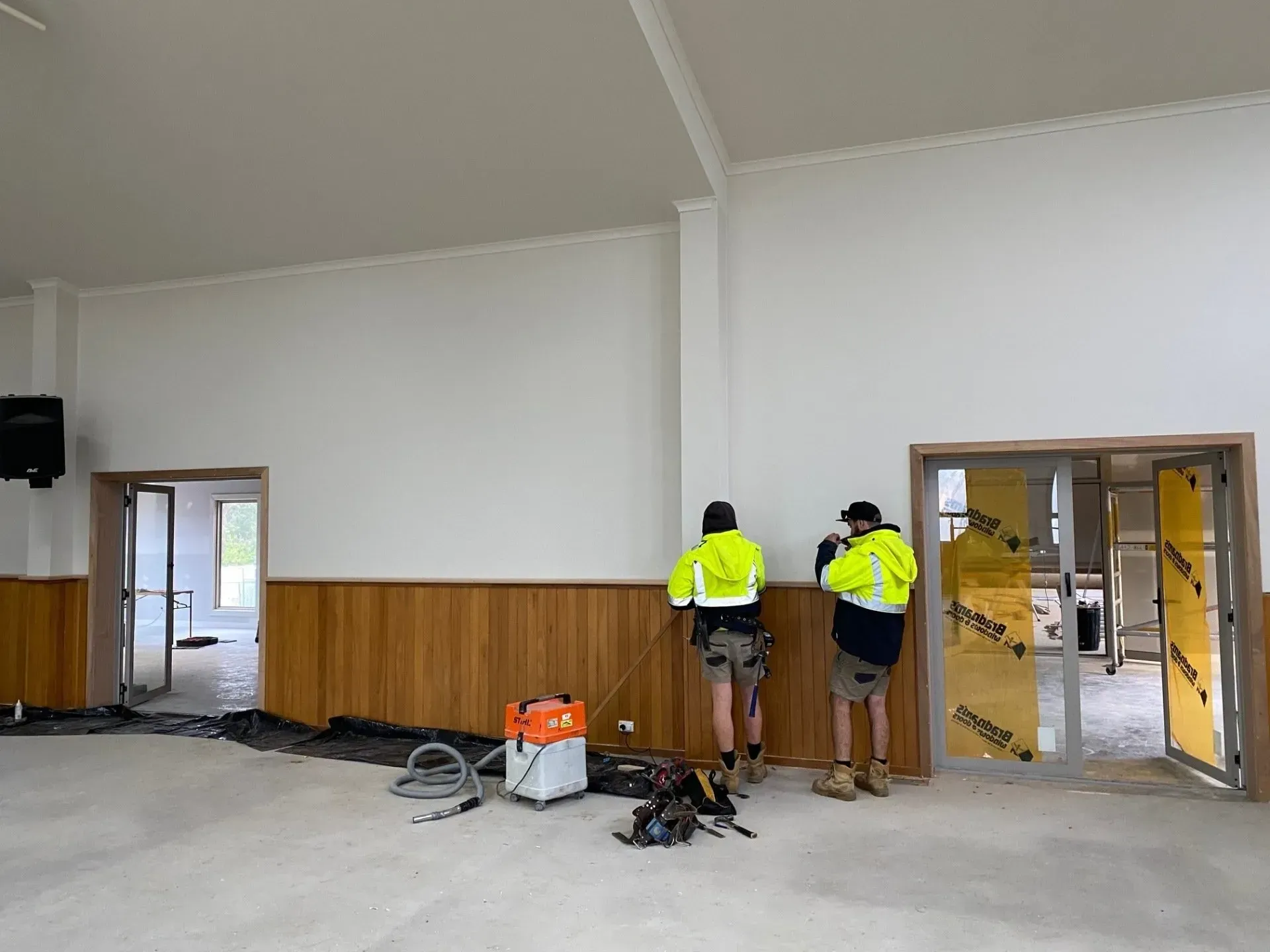 Two workers in neon vests stand in a large room with white walls during renovation.