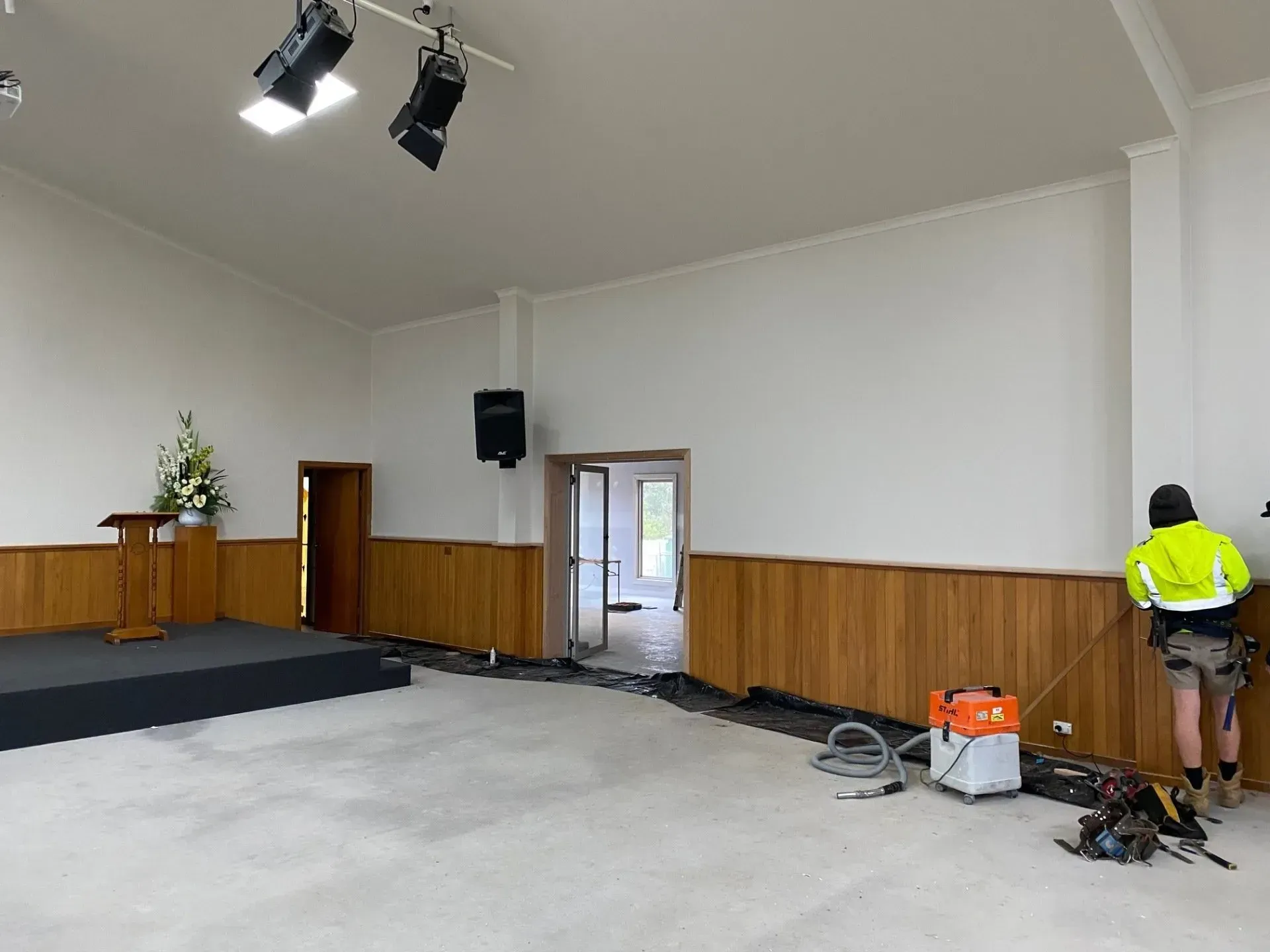 Empty community hall with stage, wooden wall paneling, and two workers on the right near equipment.