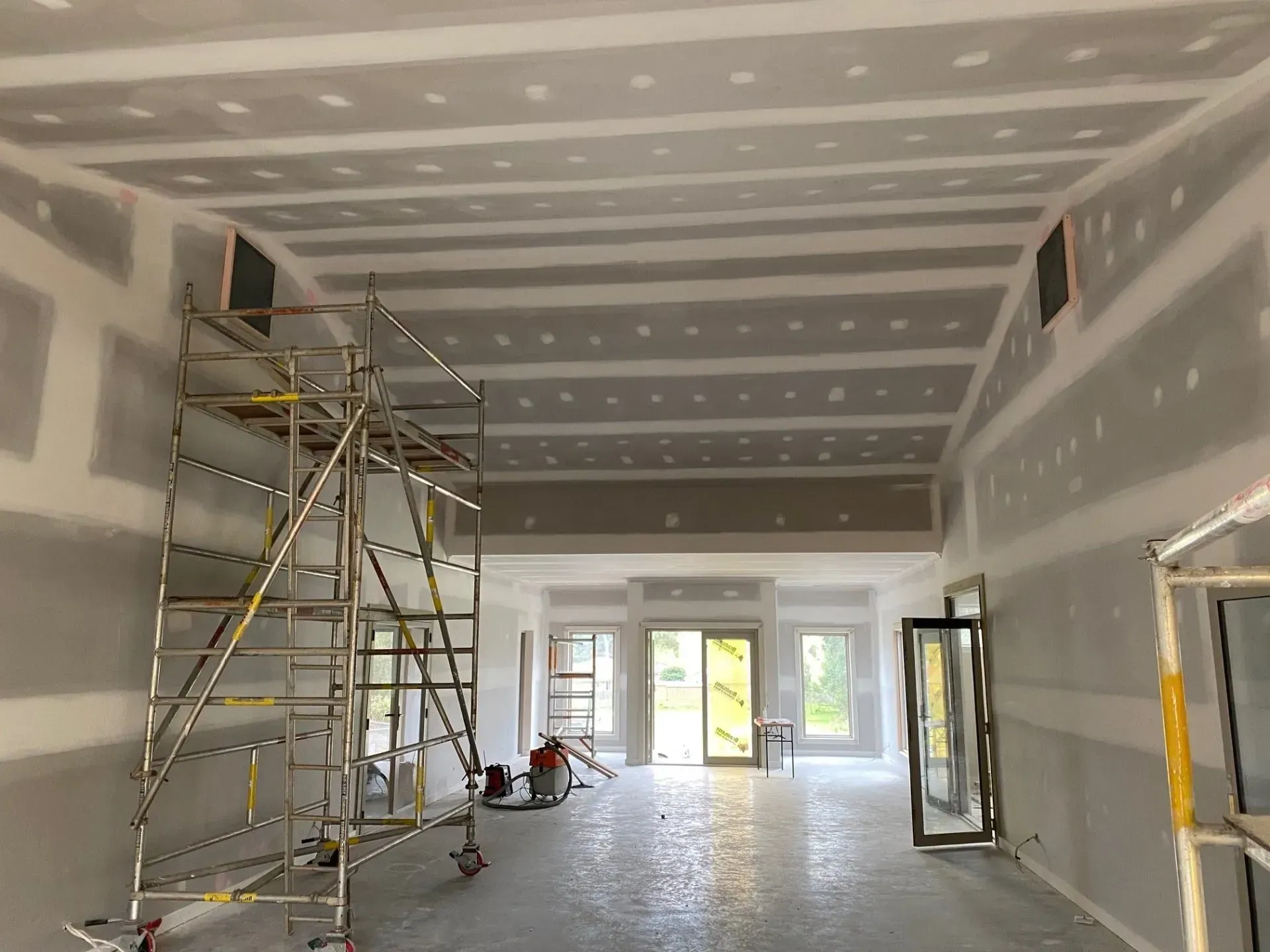 Interior under construction with drywall, scaffolding, and unfinished ceiling in a long room.