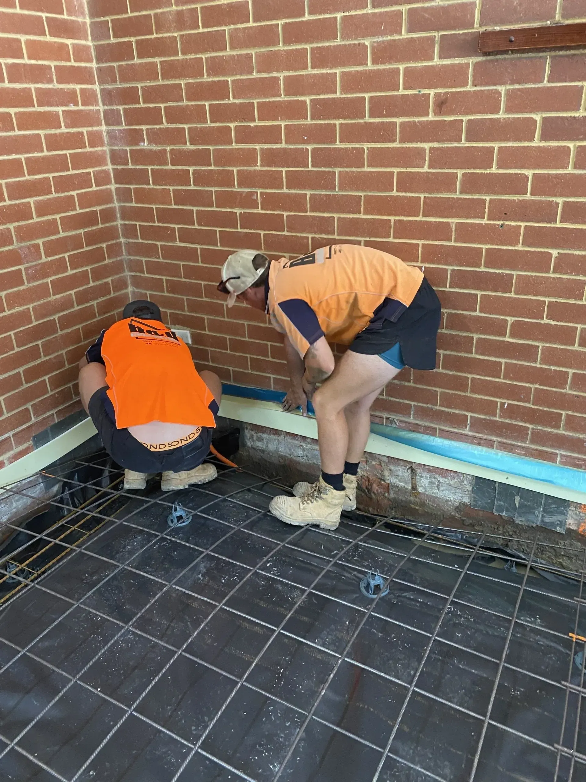 Two workers in orange vests crouch by a brick wall, inspecting or repairing the corner near the floor.