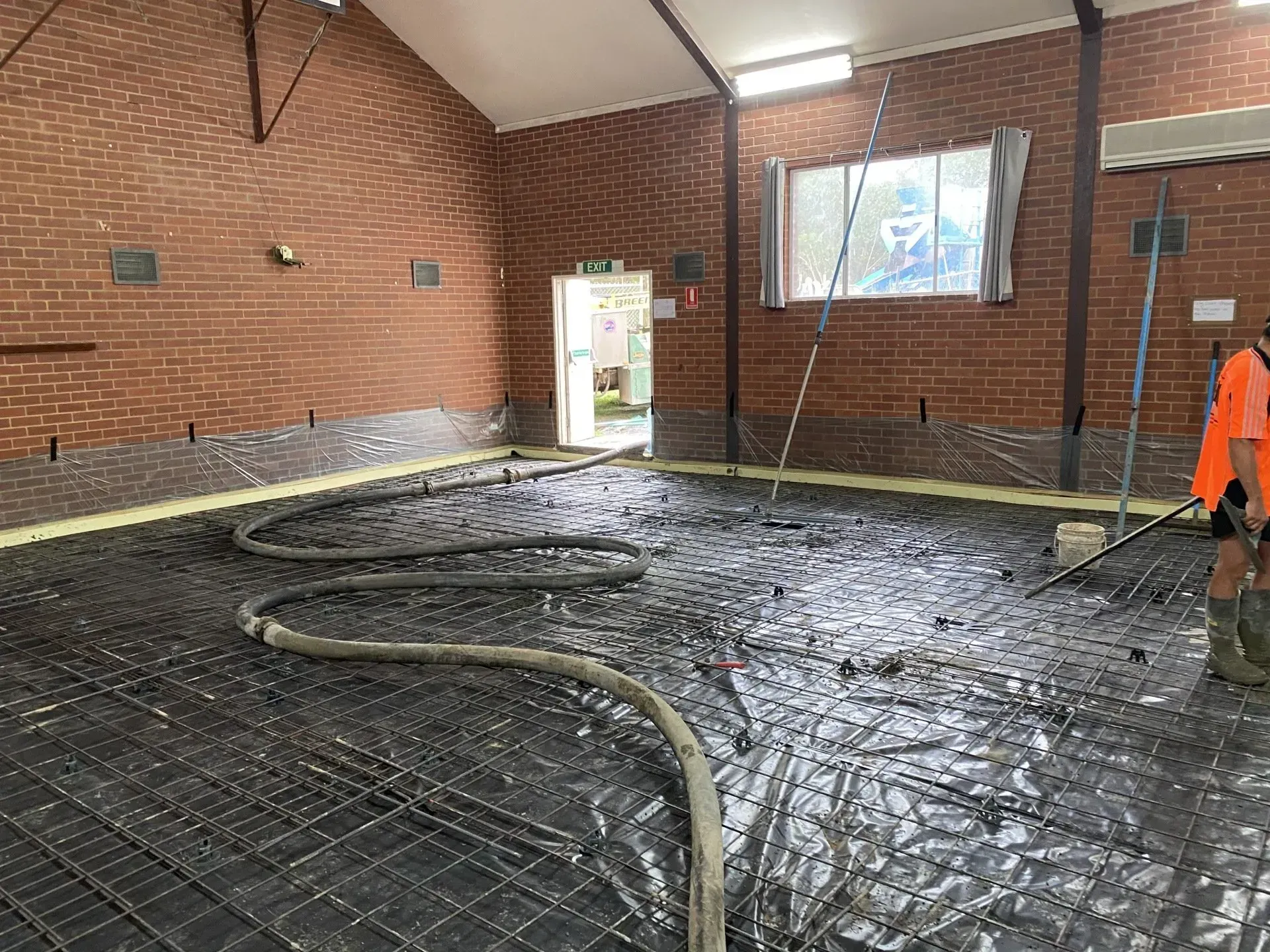 Mud-covered indoor construction floor with hoses, brick walls, and a worker in orange at right.
