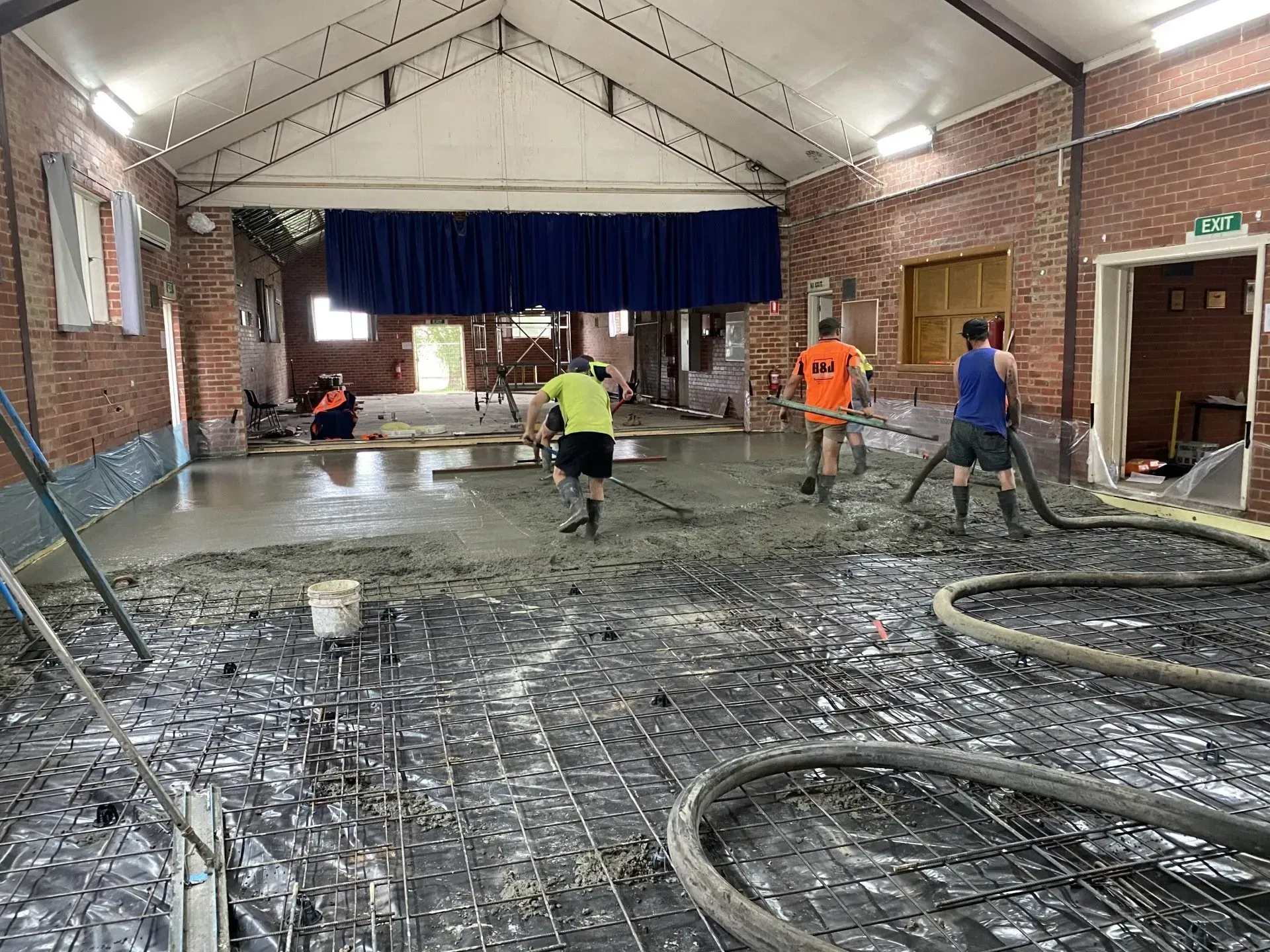 Workers smoothing wet concrete in a long indoor hall with hoses and a blue curtain stage at the far end