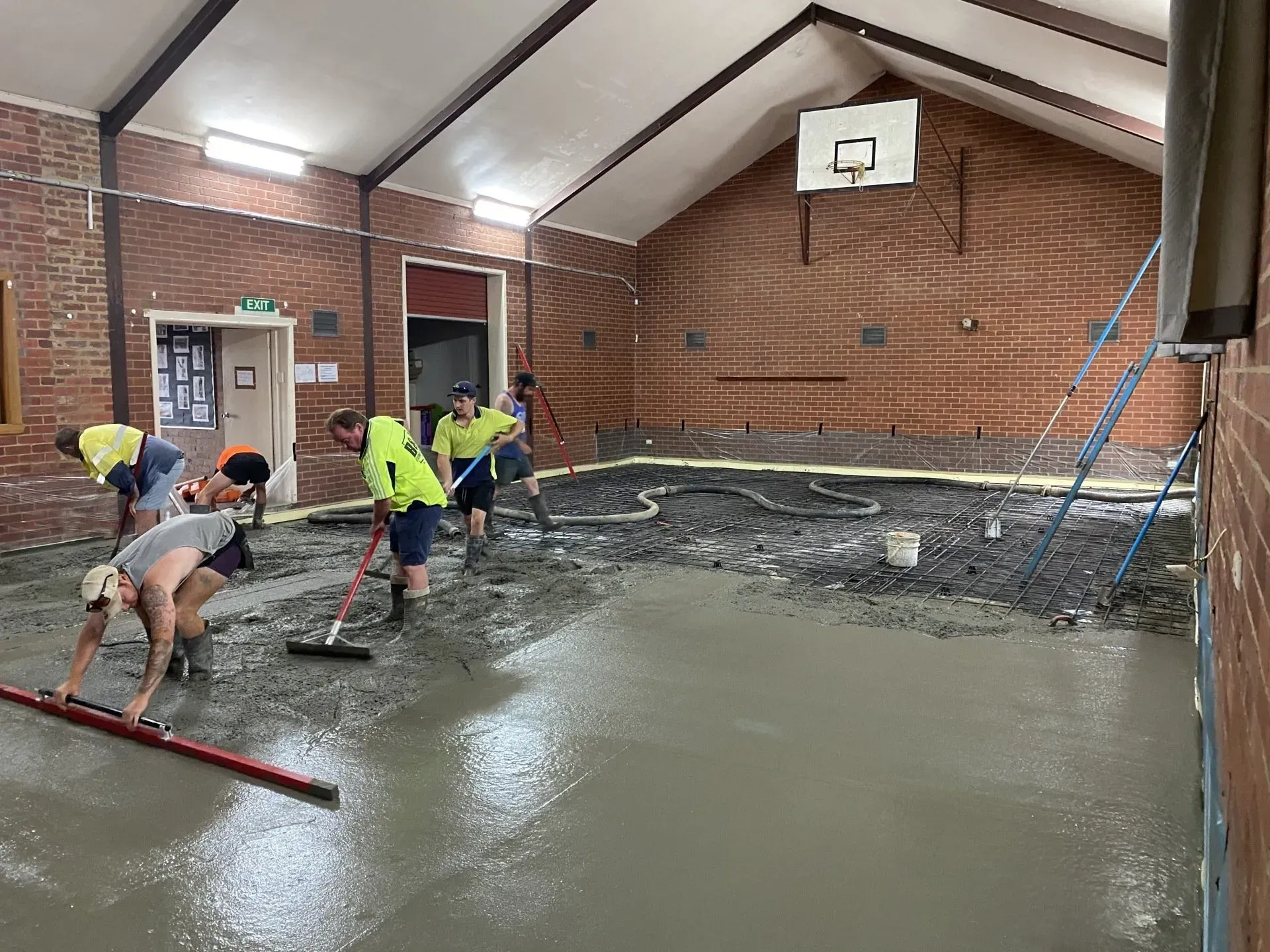 Workers spread and level wet concrete in an indoor gym with a basketball hoop.
