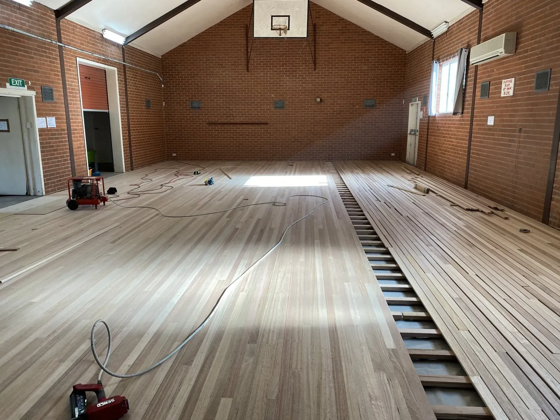 Empty brick gymnasium with a partially installed wooden floor and exposed joists on one side.