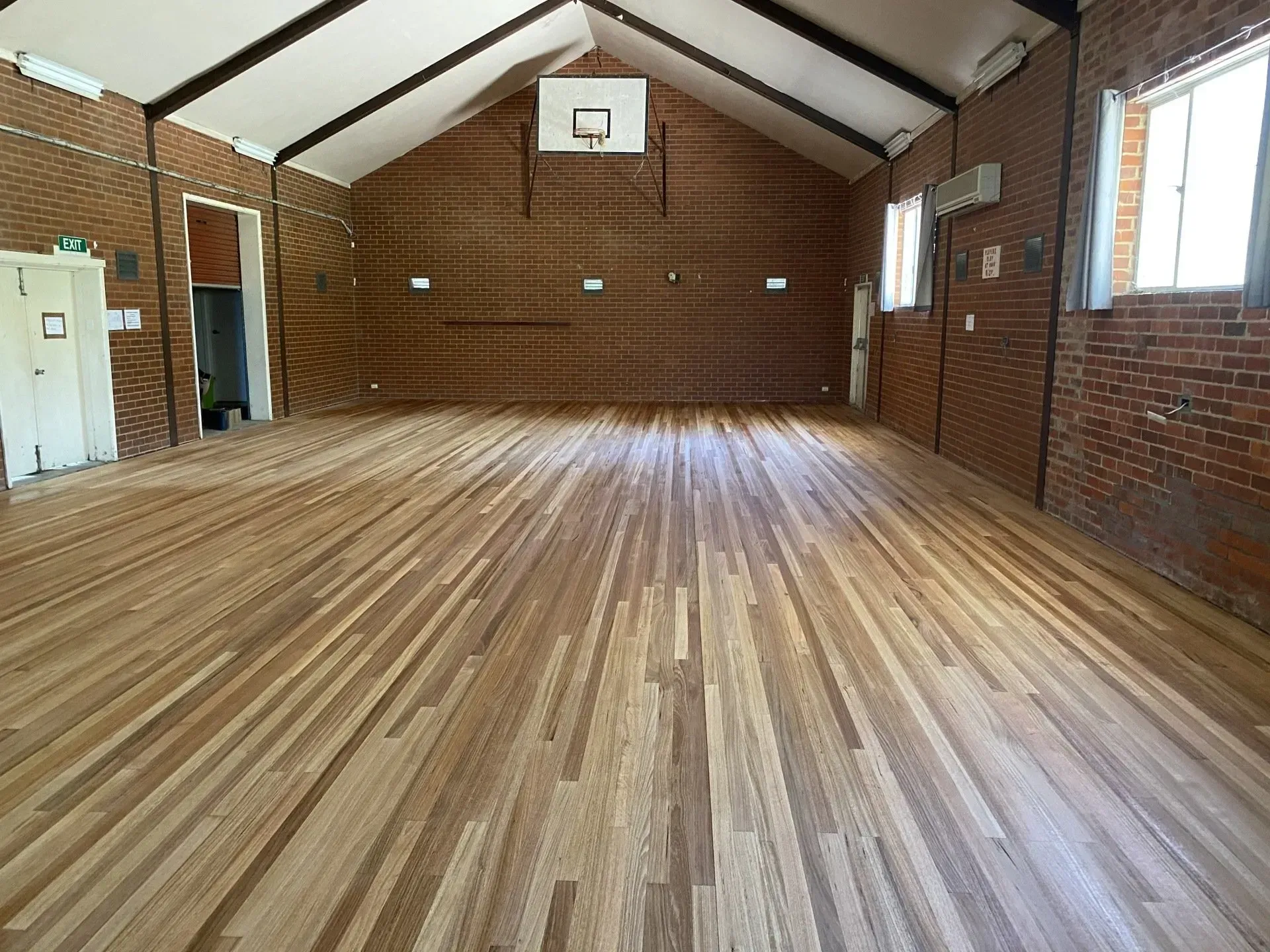 Empty brick-walled hall with wood floor, high vaulted ceiling, and a basketball hoop at the far end