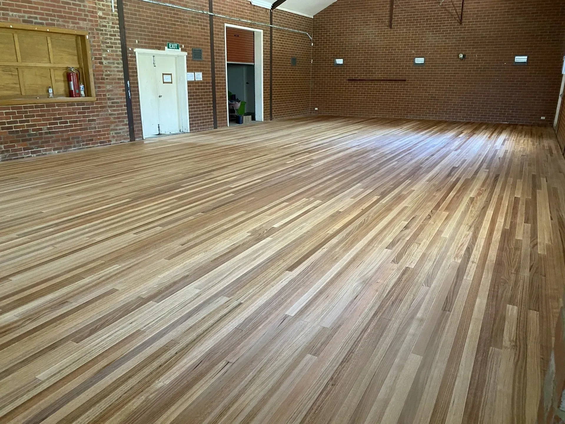 Empty wooden gymnasium floor in a brick-walled hall with sunlight streaming in from the right.