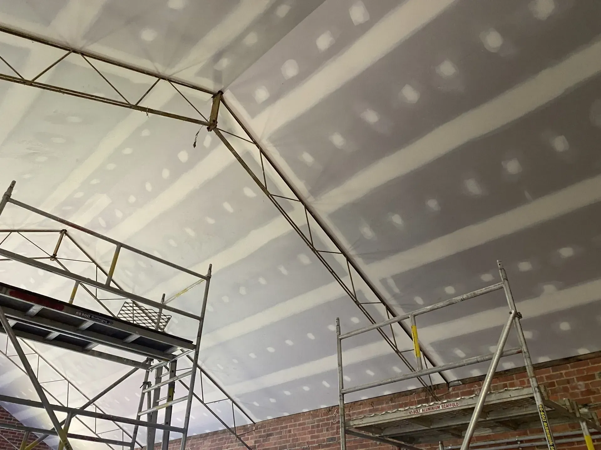 Scaffolding inside a building under construction, with drywall ceiling and brick walls visible.